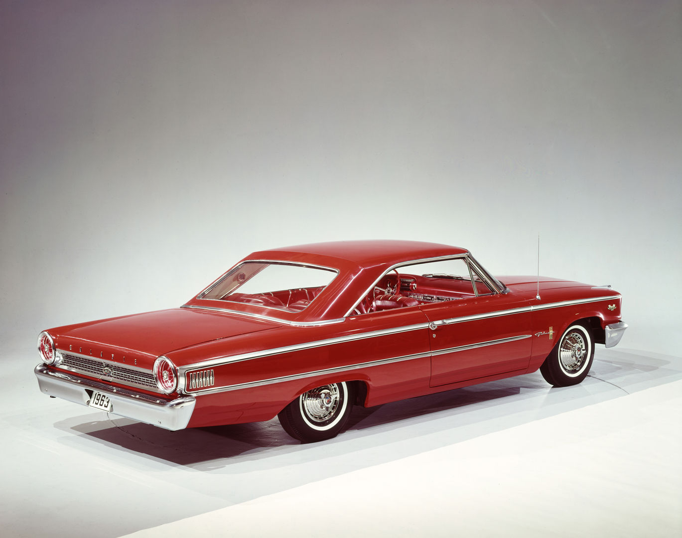 Red 1963 Ford Galaxie 500XL fastback hardtop in a studio, high rear three-quarter view, showing the sloping roof and red bucket-seat interior