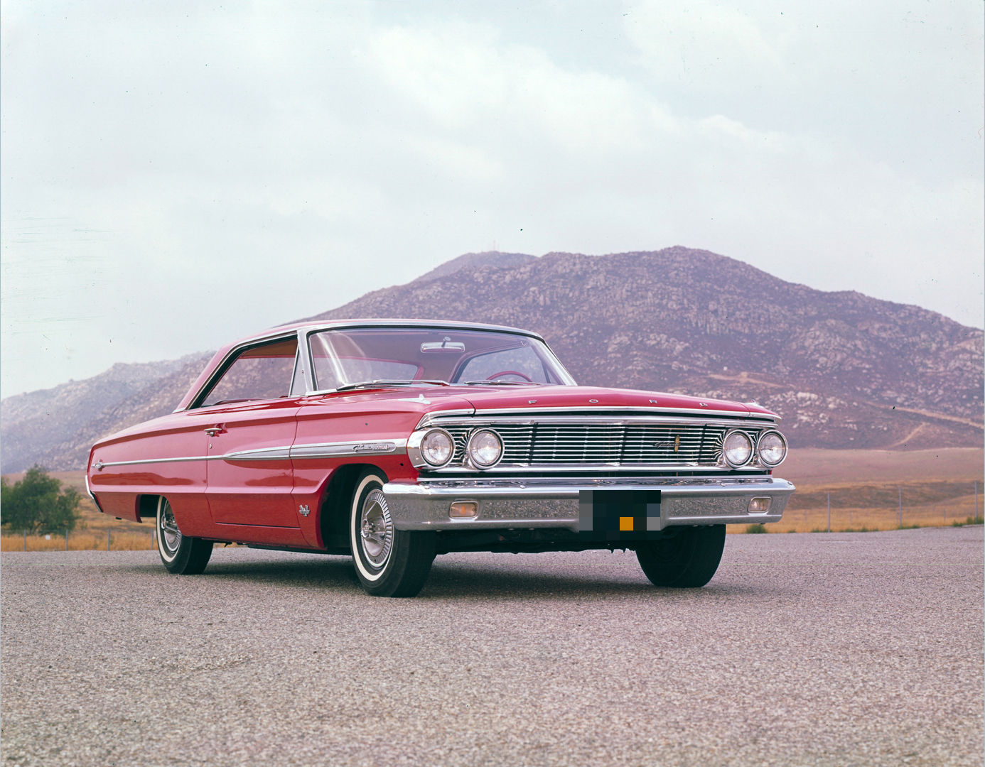 Red 1964 Ford Galaxie 500XL two-door hardtop, low front three-quarter view on a paved lot with a rocky hill behind, showing the quad-headlight grille
