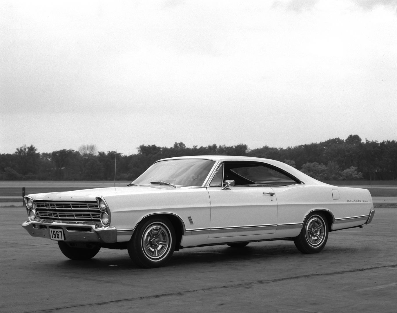 Black-and-white photo of a light-colored 1967 Ford Galaxie 500 fastback hardtop on an airfield apron, front three-quarter view, showing the stacked headlights