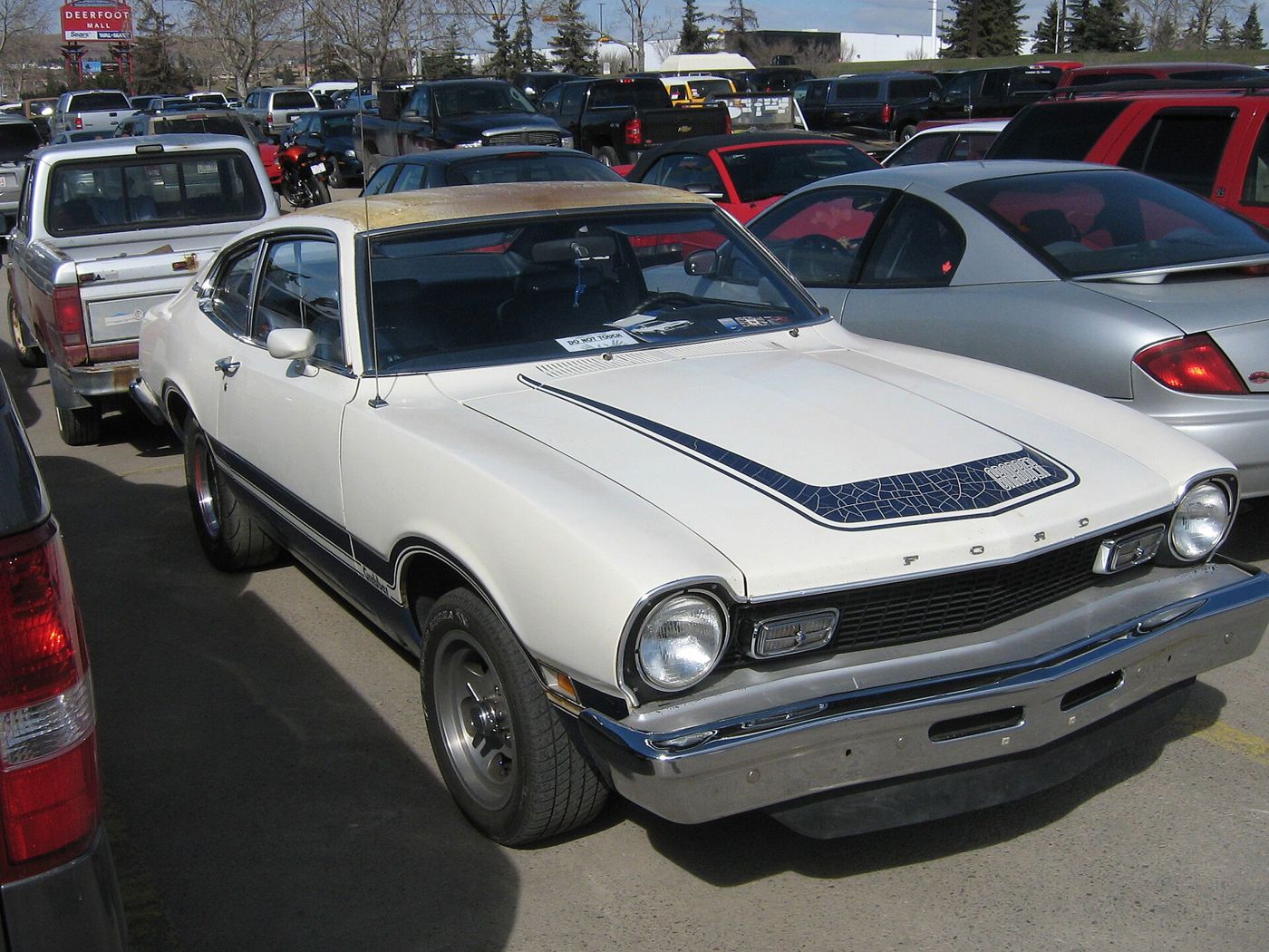 White Ford Maverick Grabber with blue hood and side graphics and the larger front bumper, front three-quarter view in a crowded parking lot