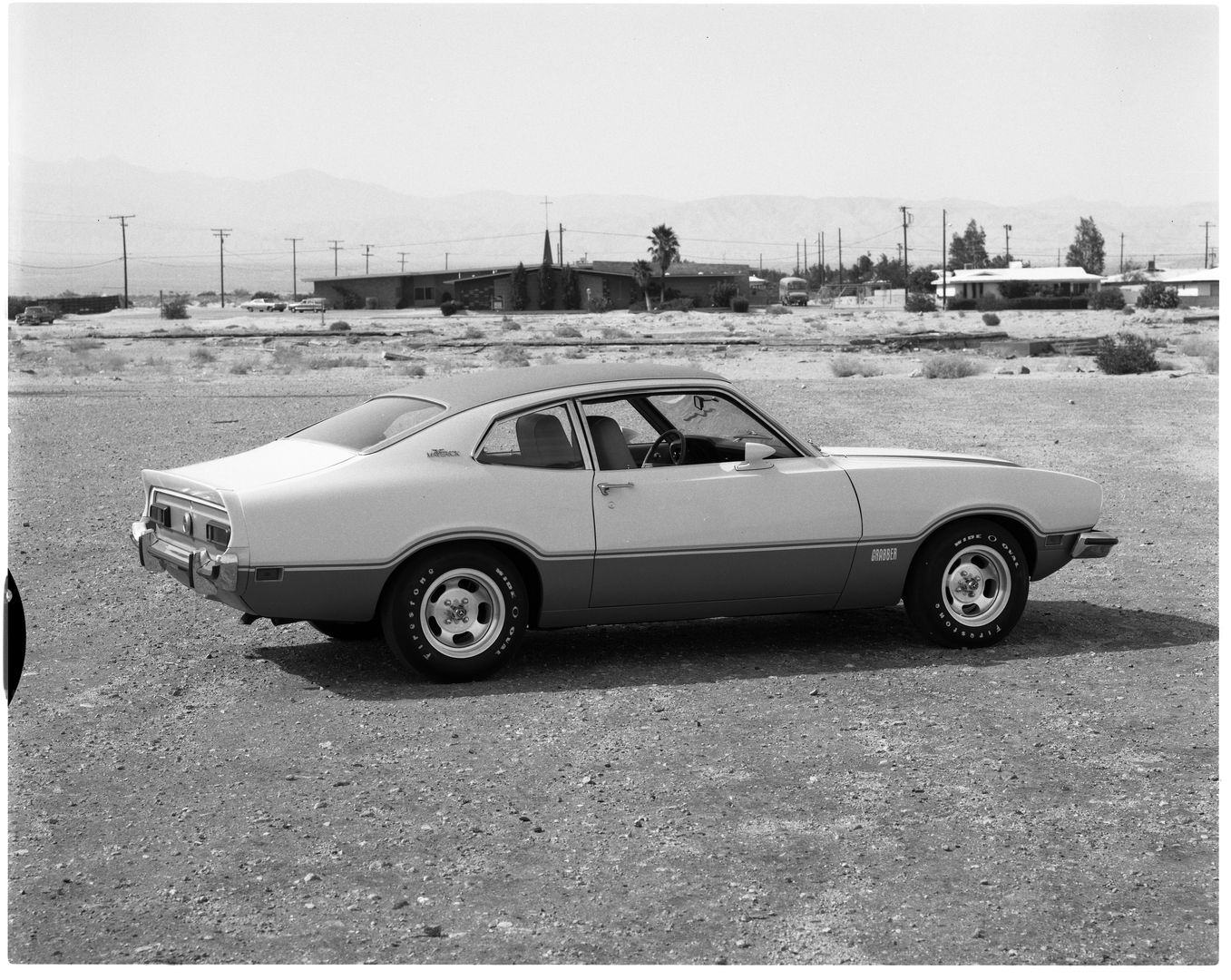 Black-and-white photo of a two-tone 1973 Ford Maverick Grabber seen from the rear three-quarter on a desert lot with houses and power poles behind