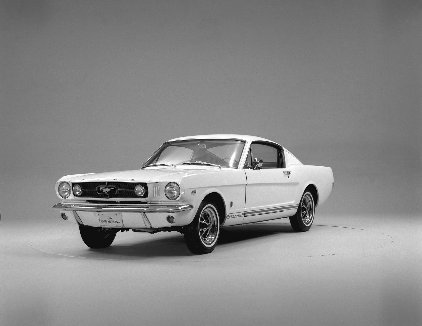 Black-and-white studio photo of a white 1965 Ford Mustang GT fastback, front three-quarter view, with grille-mounted fog lamps, GT rocker stripe and styled wheels