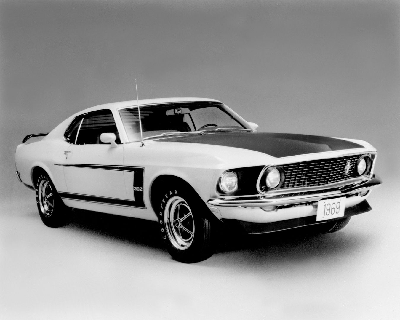 Black-and-white studio photo of a white 1969 Ford Mustang Boss 302 fastback, front three-quarter view, showing the side C-stripe with 302 lettering, blacked-out hood and Magnum 500 wheels