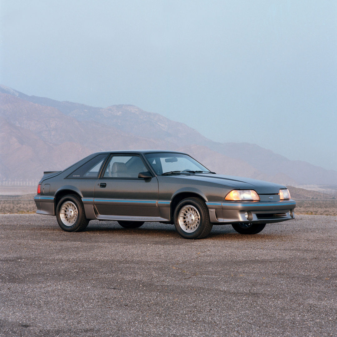 Grey 1987 Ford Mustang GT hatchback with body-color ground effects and turbine-style wheels, front three-quarter view on a desert lot below mountains