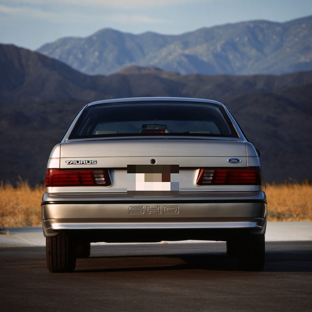 Silver 1989 Ford Taurus SHO seen straight from behind with mountains beyond, showing the taillight band and SHO lettering molded into the rear bumper