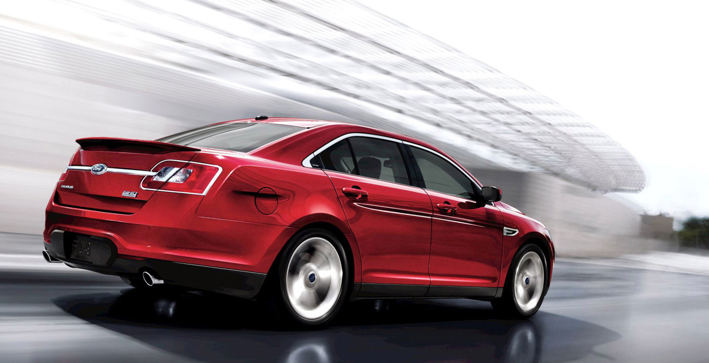 Red 2011 Ford Taurus SHO driving past a curved modern building, rear three-quarter view, showing the decklid spoiler and SHO badge