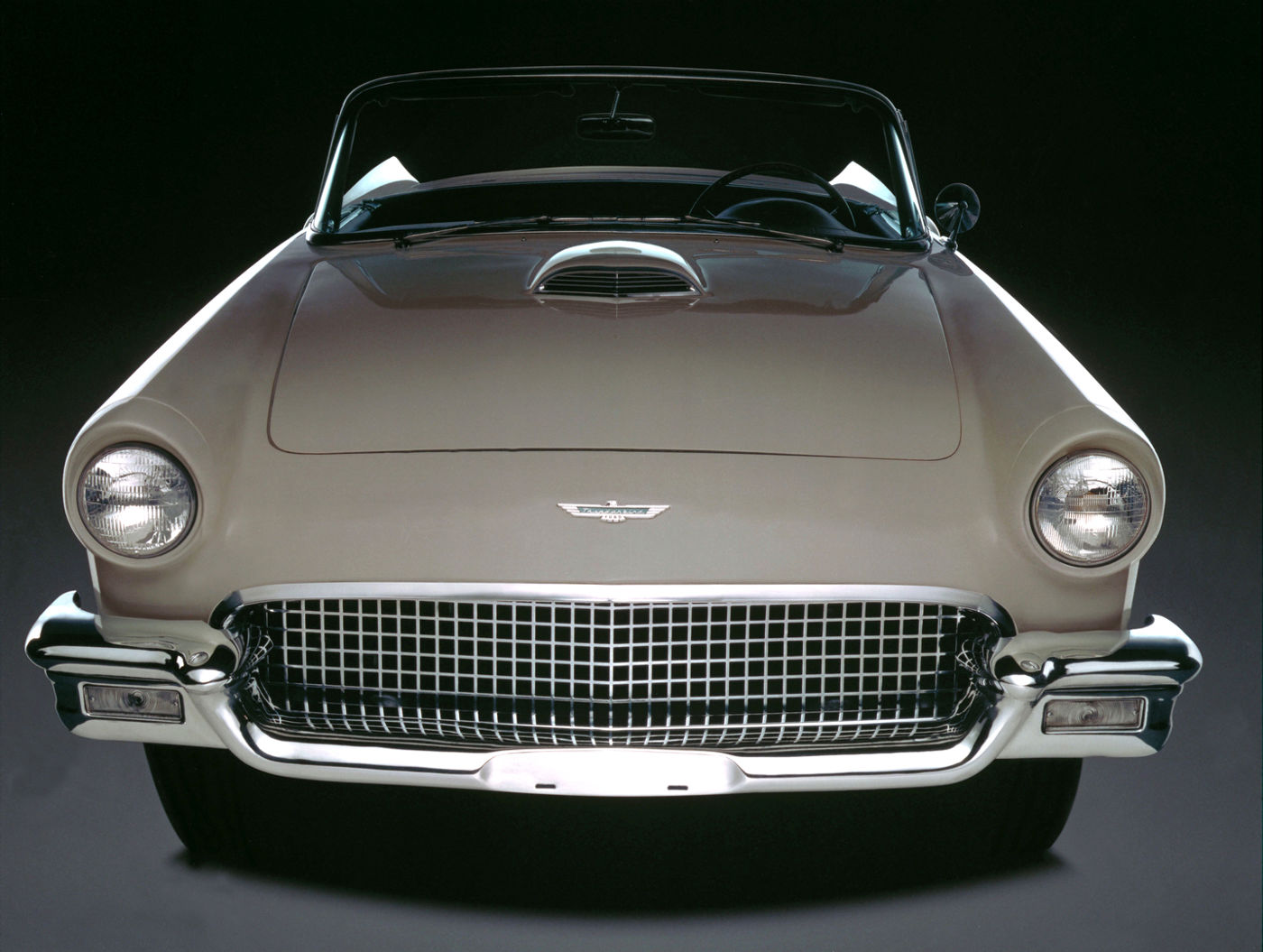 Beige 1957 Ford Thunderbird seen head-on on a black studio backdrop, showing the hood scoop, round headlamps and the wide eggcrate grille and bumper