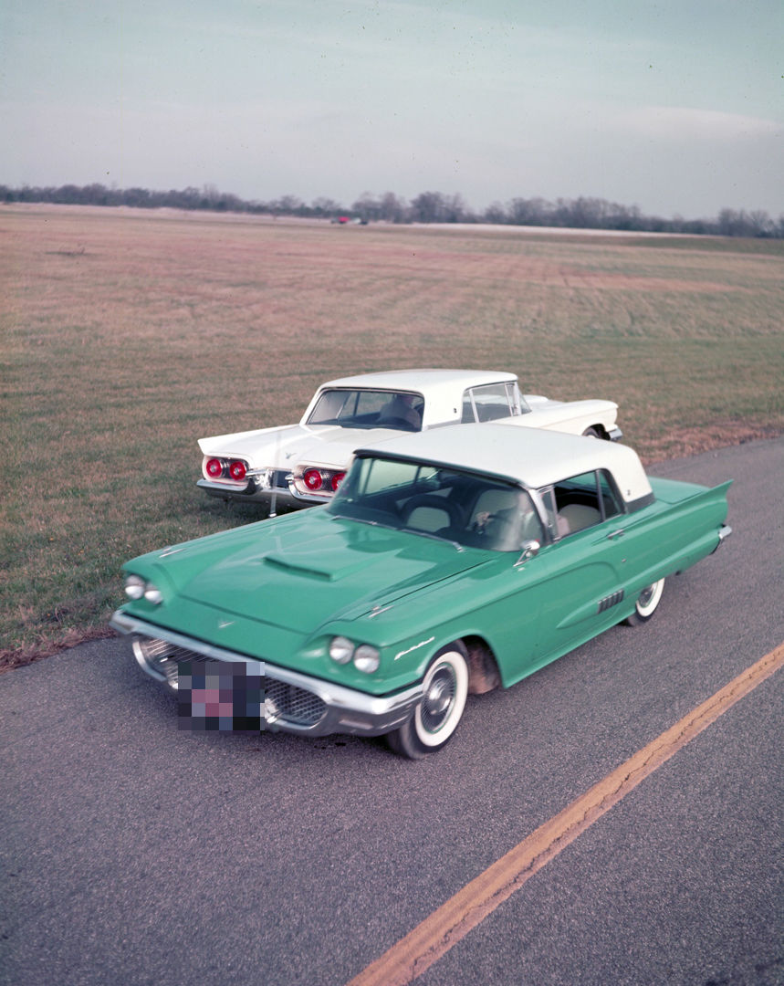 Two 1958 Ford Thunderbird hardtops, one turquoise and one white, on a test-track road, the turquoise car in front three-quarter view