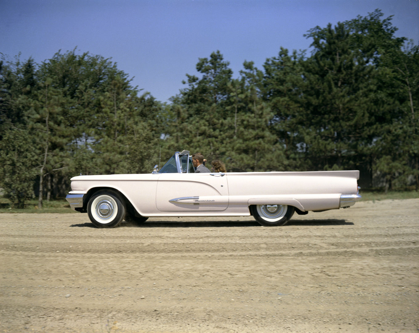 White 1959 Ford Thunderbird convertible in full side profile on a dirt road in front of pine trees, with two occupants