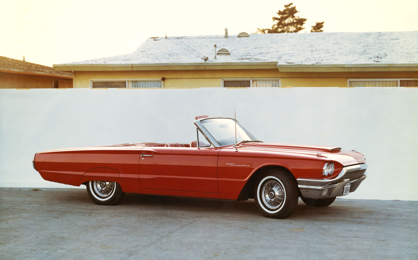 Red 1964 Ford Thunderbird convertible with the top down, front three-quarter side view in front of a low house