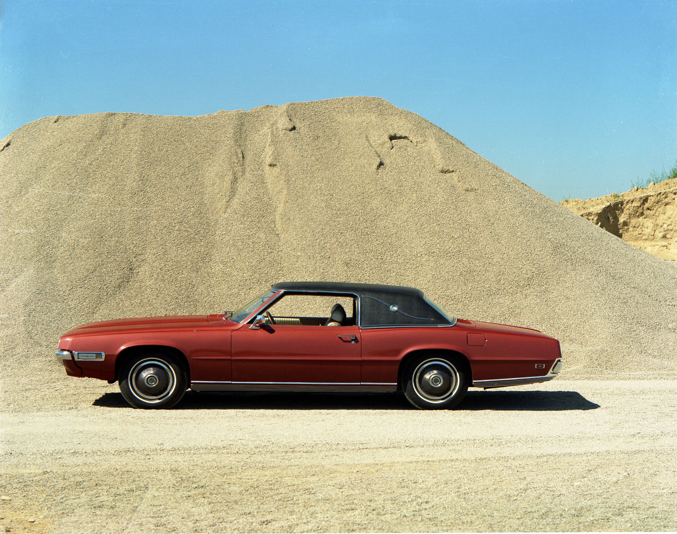 Red 1969 Ford Thunderbird Landau two-door with a black vinyl roof in side profile in front of a gravel pile