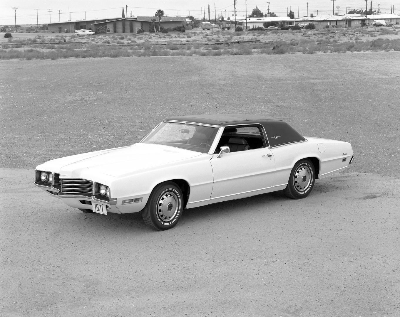 Black-and-white photo of a white 1971 Ford Thunderbird two-door with a dark roof on a desert lot, front three-quarter view showing the projecting beak grille