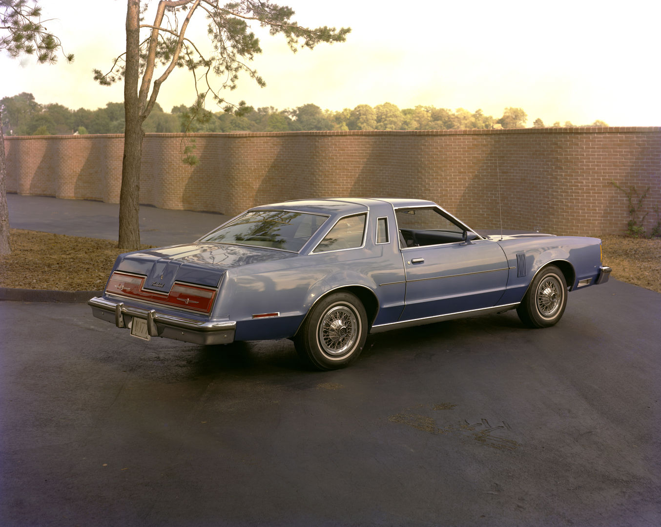 Light blue 1977 Ford Thunderbird with a wrapover roof band, rear three-quarter view on a paved lot by a brick wall