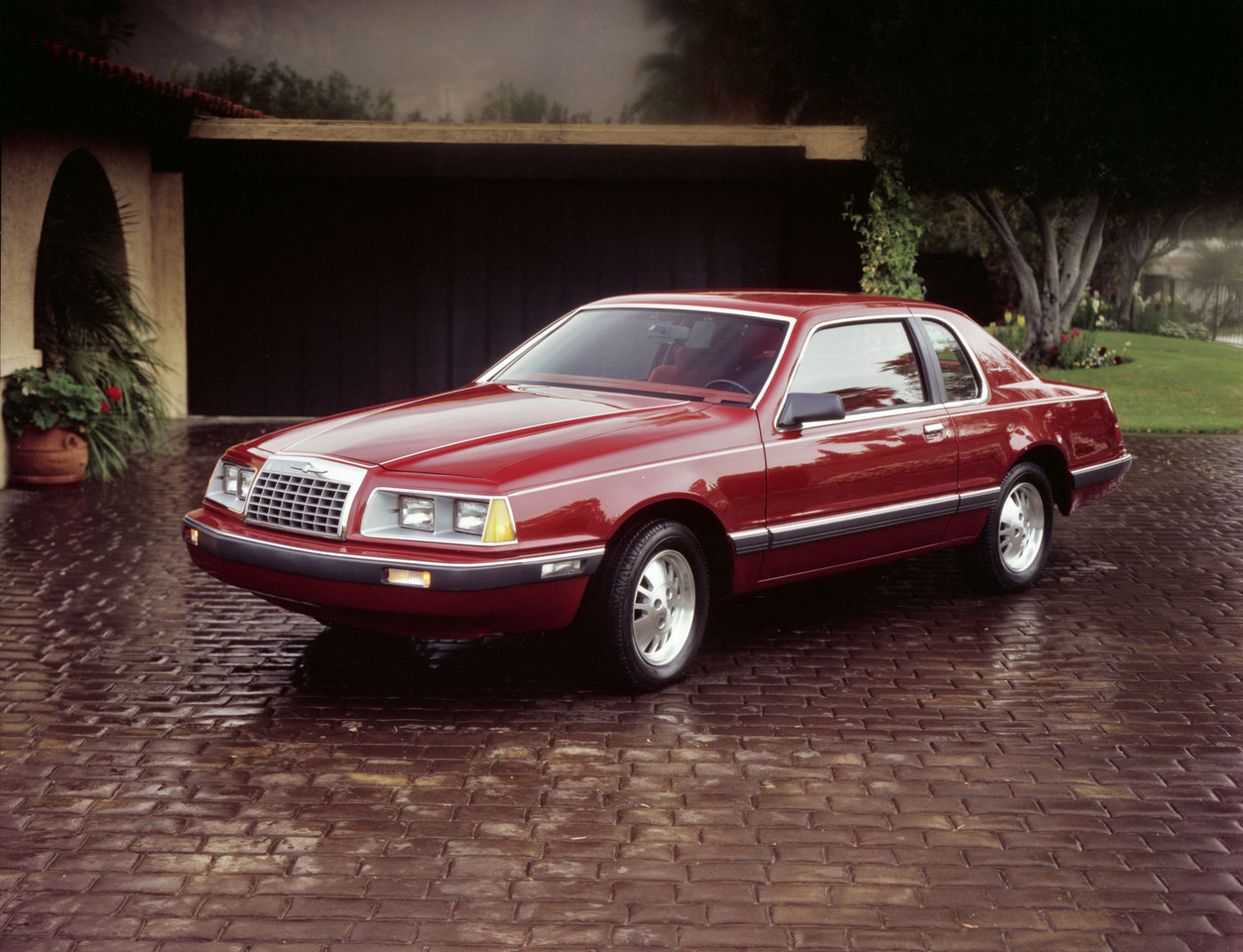 Red 1983 Ford Thunderbird coupe on wet cobblestones before a garage, front three-quarter view