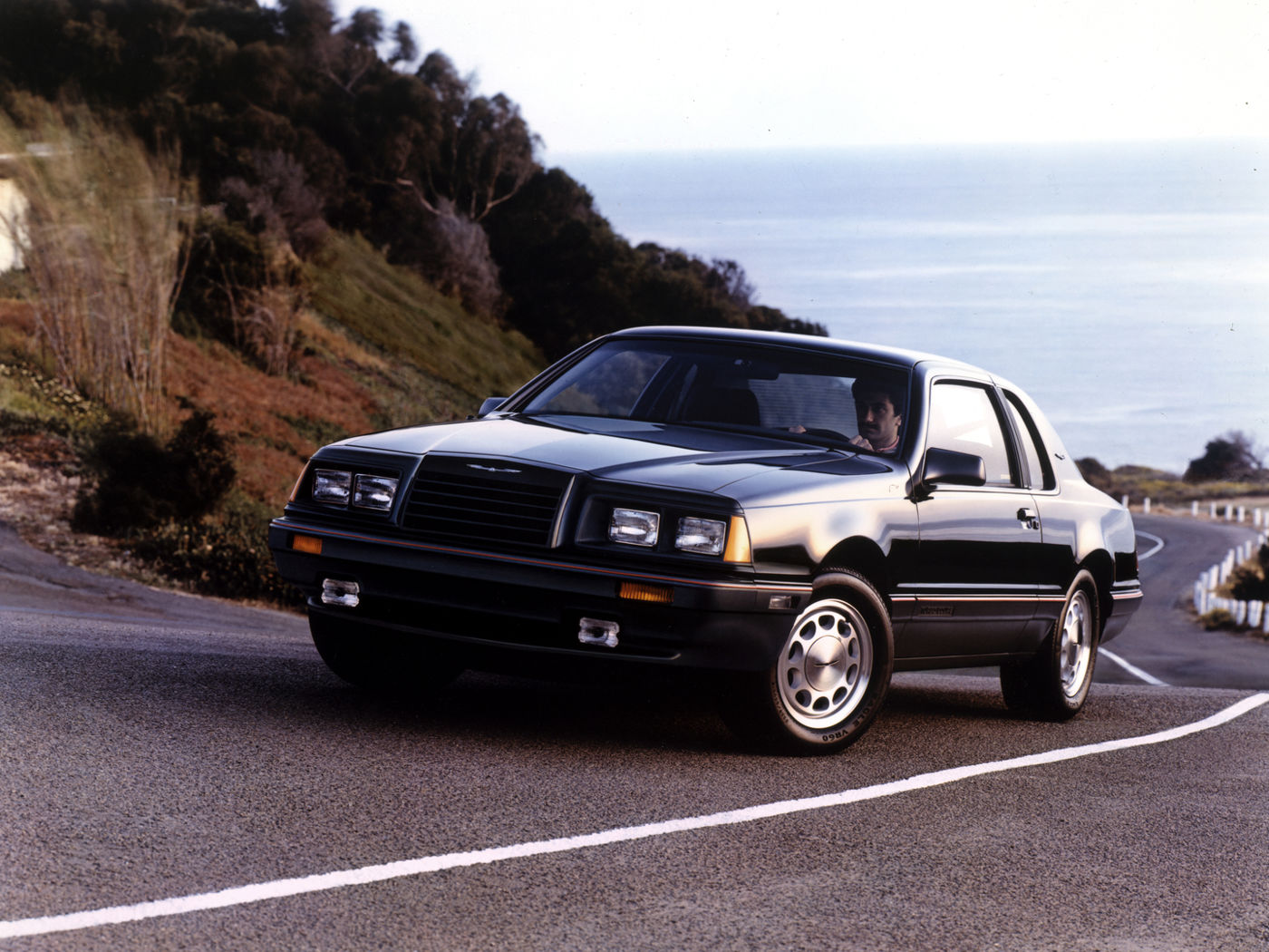 Black 1986 Ford Thunderbird Turbo Coupe on a coastal road, front three-quarter view