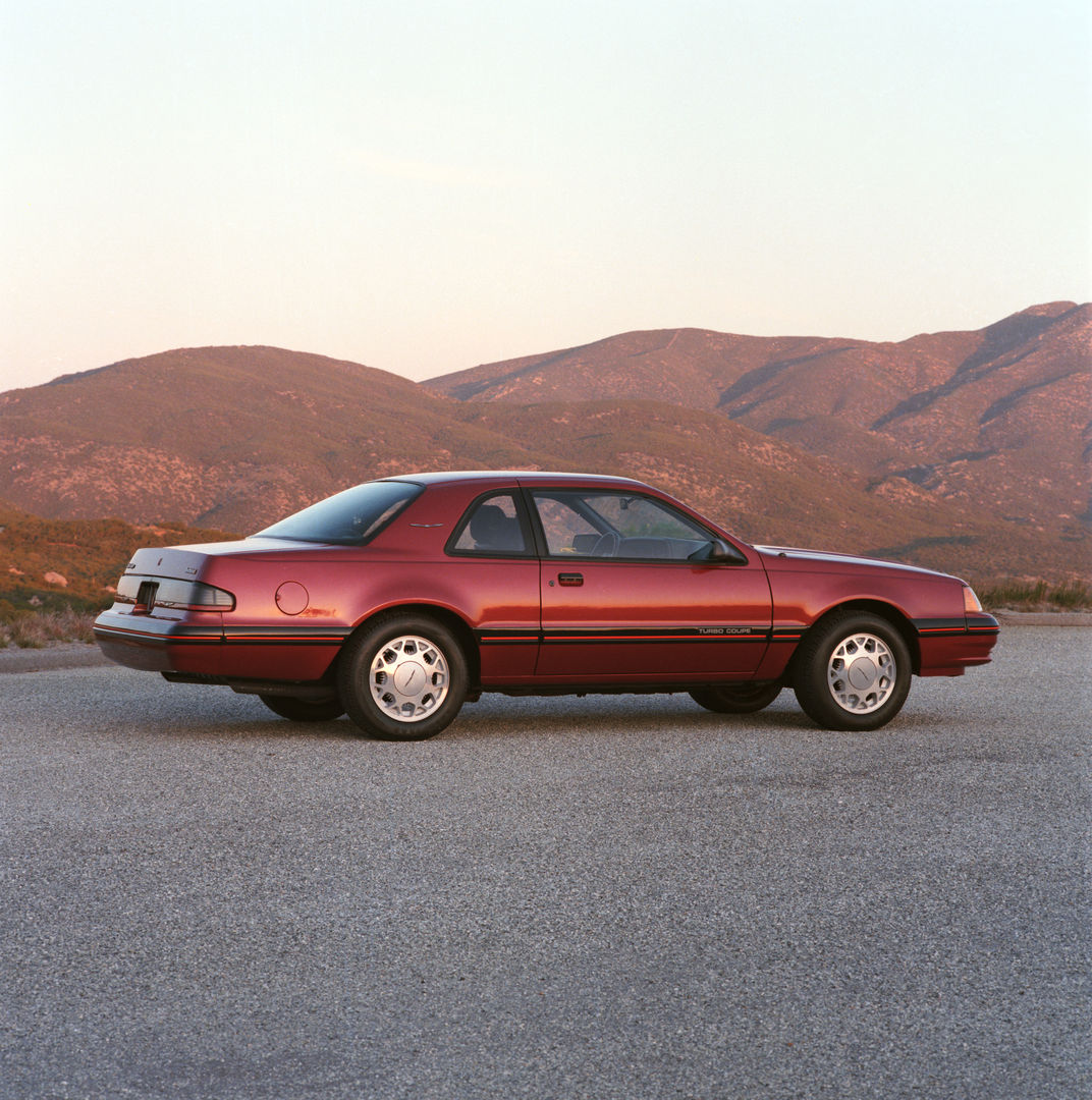 Dark red 1987 Ford Thunderbird Turbo Coupe on a paved lot below hills at dusk, rear three-quarter view