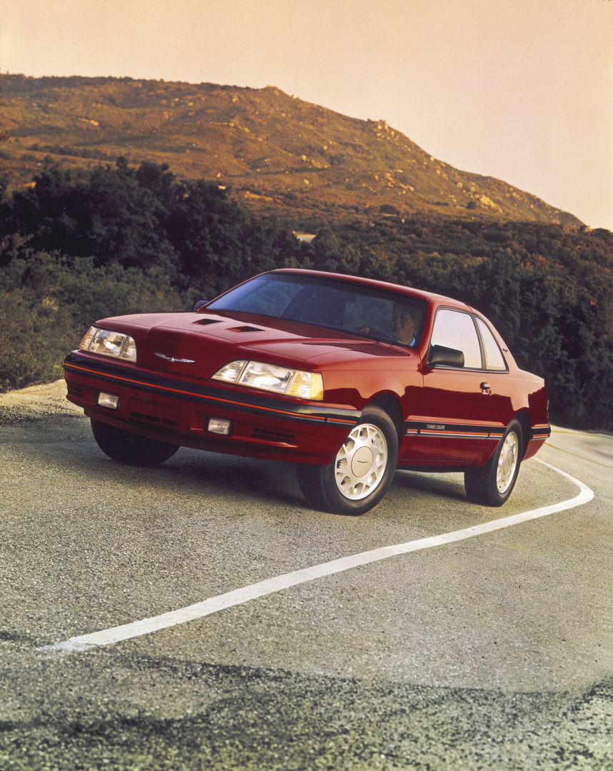 Red 1988 Ford Thunderbird Turbo Coupe on a winding road, front three-quarter view with the twin hood scoops