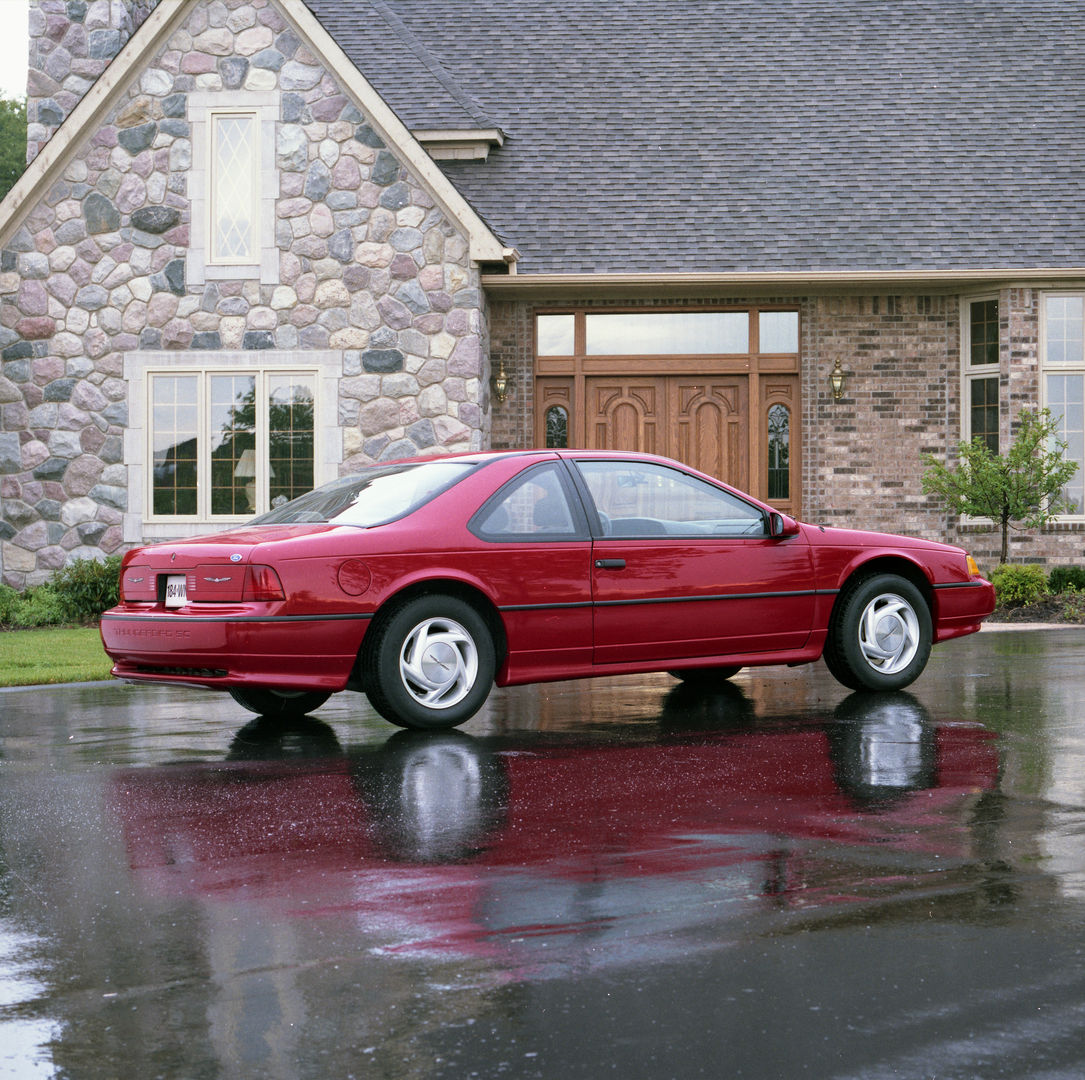 Red 1989 Ford Thunderbird Super Coupe on a wet driveway before a stone house, rear three-quarter view