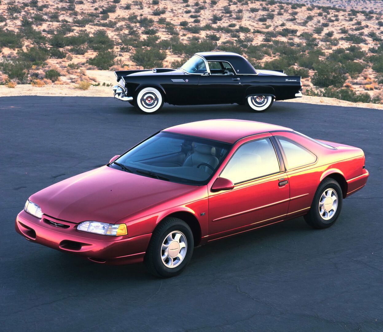 Red 1995 Ford Thunderbird coupe in the foreground and a black 1955 Thunderbird with a hardtop behind it on a desert road