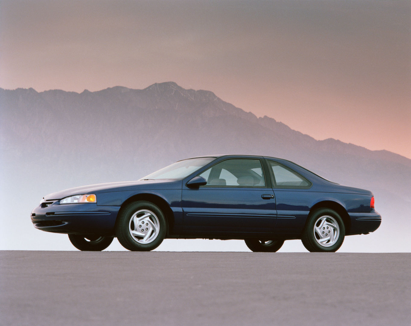 Dark blue 1996 Ford Thunderbird LX coupe in side profile on pavement before pink-lit mountains