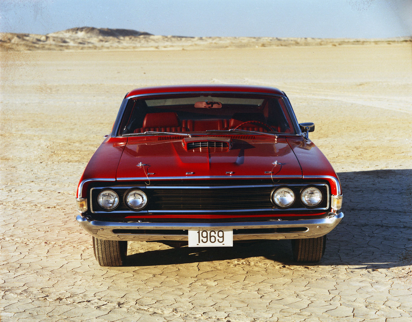 Red 1969 Ford Torino Cobra seen head-on on a dry lake bed, showing the hood scoop, hood pins and quad headlights in a black grille