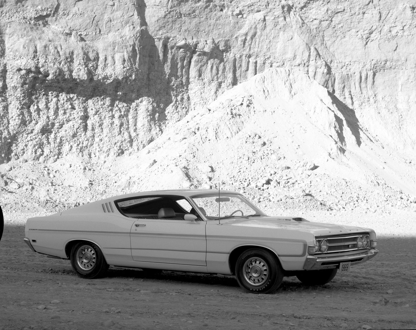 Black-and-white photo of a light-colored 1969 Ford Torino GT fastback parked below a quarry wall, side and front three-quarter view, with a hood scoop and GT grille badge
