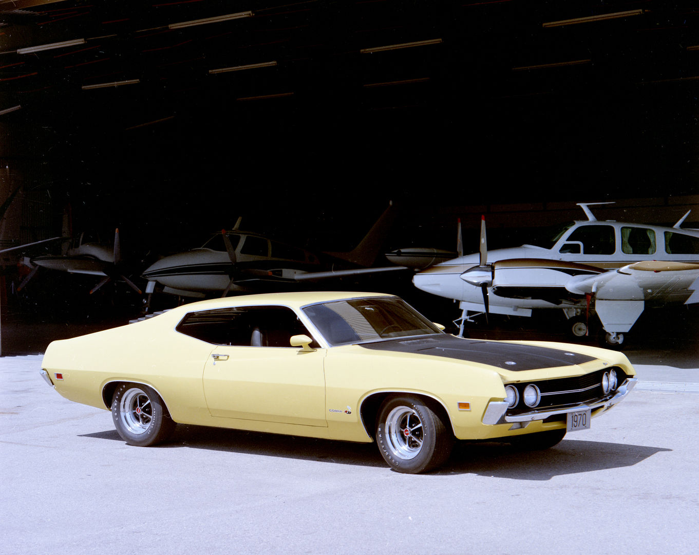 Pale yellow 1970 Ford Torino Cobra fastback with a flat black hood and Magnum 500 wheels, front three-quarter view in front of an aircraft hangar with light planes inside