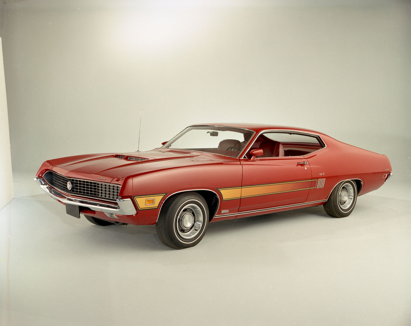 Red 1970 Ford Torino GT fastback in a studio, front three-quarter view, with a gold side stripe, hood scoop and a 429 emblem on the rocker