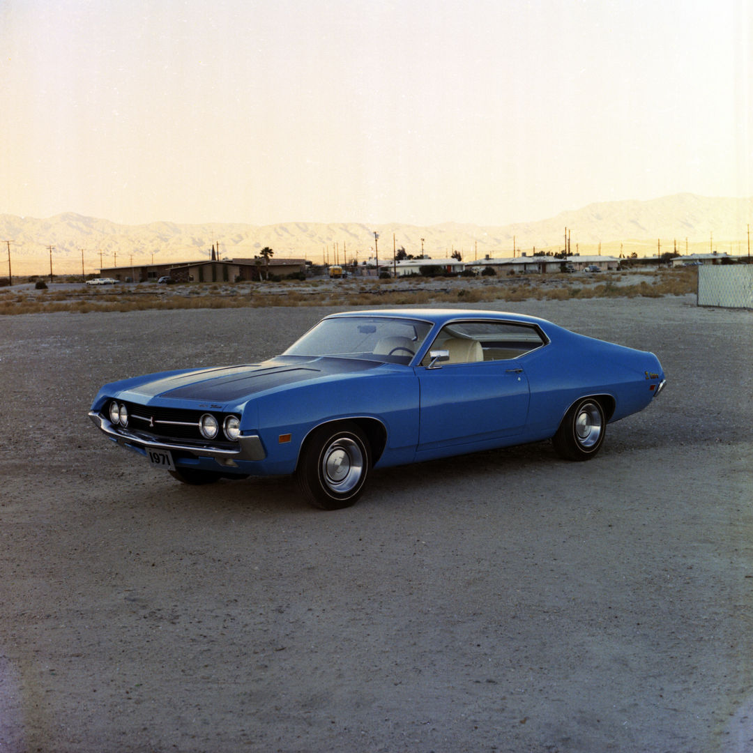 Blue 1971 Ford Torino Cobra fastback with a black hood, front three-quarter view on a gravel lot with desert mountains behind