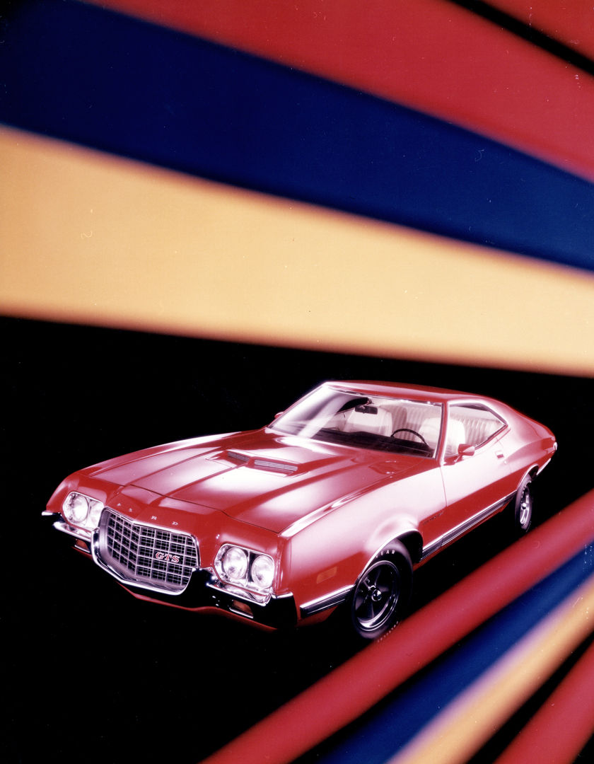 Red 1972 Ford Gran Torino Sport two-door with twin hood scoops and a large open grille, photographed against bold red, blue and yellow bands