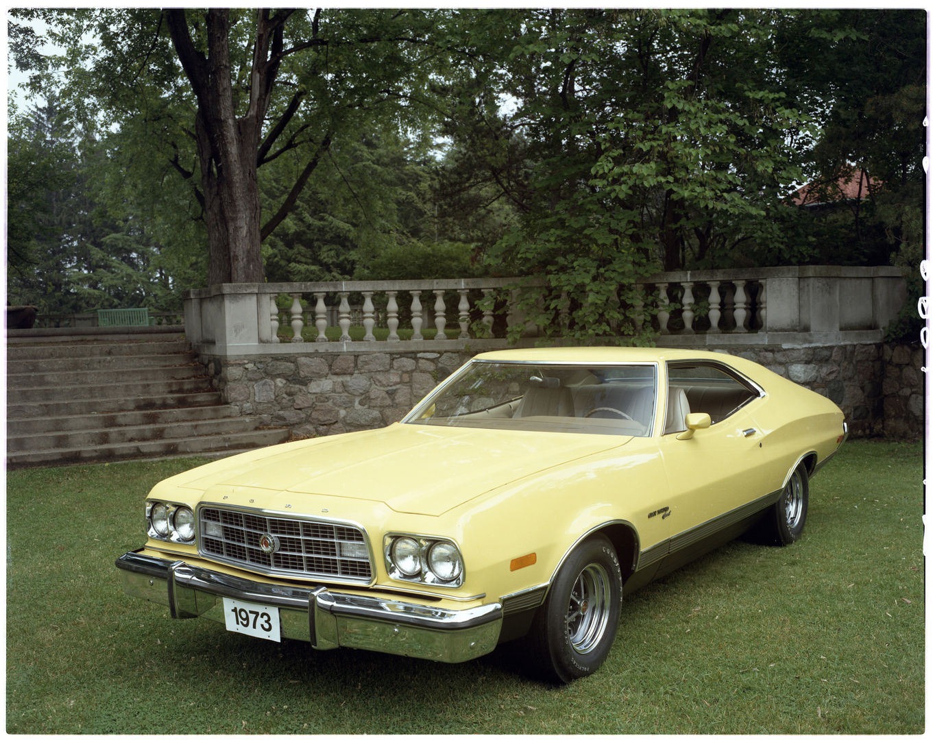 Pale yellow 1973 Ford Gran Torino Sport two-door on a lawn before a stone balustrade, front three-quarter view, with the larger 1973 bumper and Magnum 500 wheels