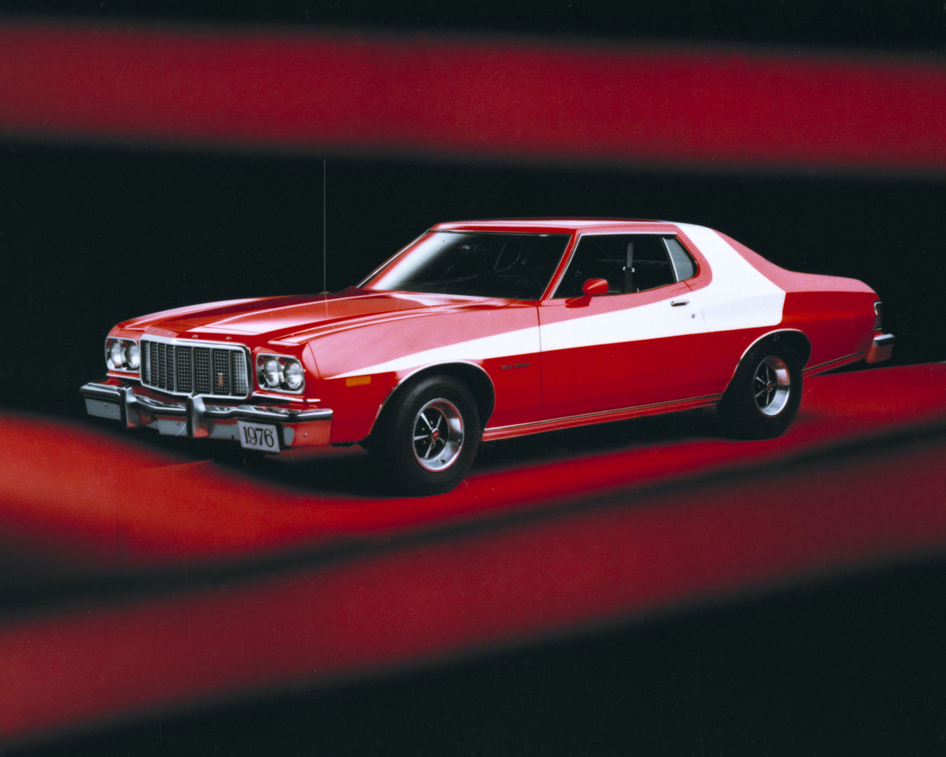 Red 1976 Ford Gran Torino two-door with a white vector stripe sweeping over the roof and slot-style wheels, front three-quarter view on a red and black backdrop