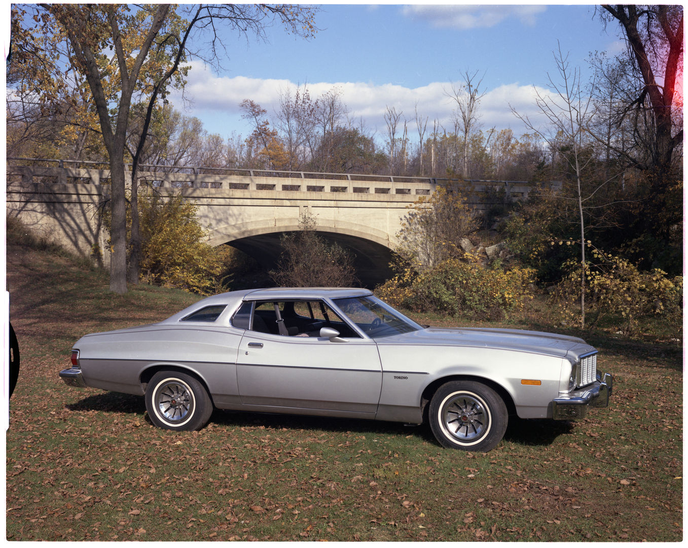 Silver 1976 Ford Torino two-door hardtop in profile on fallen leaves in front of a concrete road bridge
