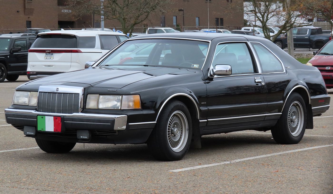Black 1986 Lincoln Mark VII LSC coupe in a parking lot, front three-quarter view, with BBS-style mesh wheels and an Italian-flag front plate