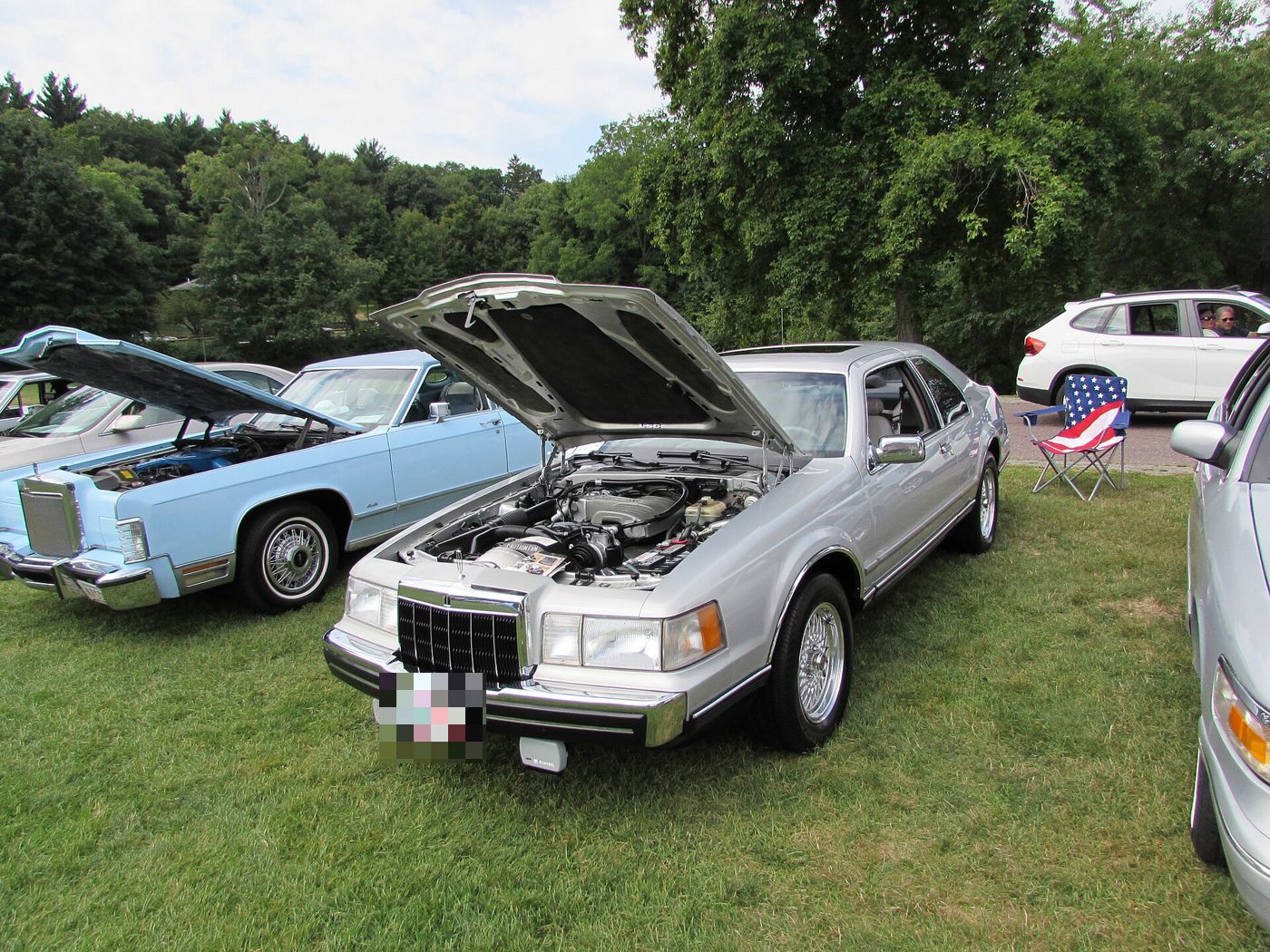 Silver 1990 Lincoln Mark VII coupe with its hood raised at an outdoor car show, front three-quarter view showing the 5.0 liter V8 engine bay