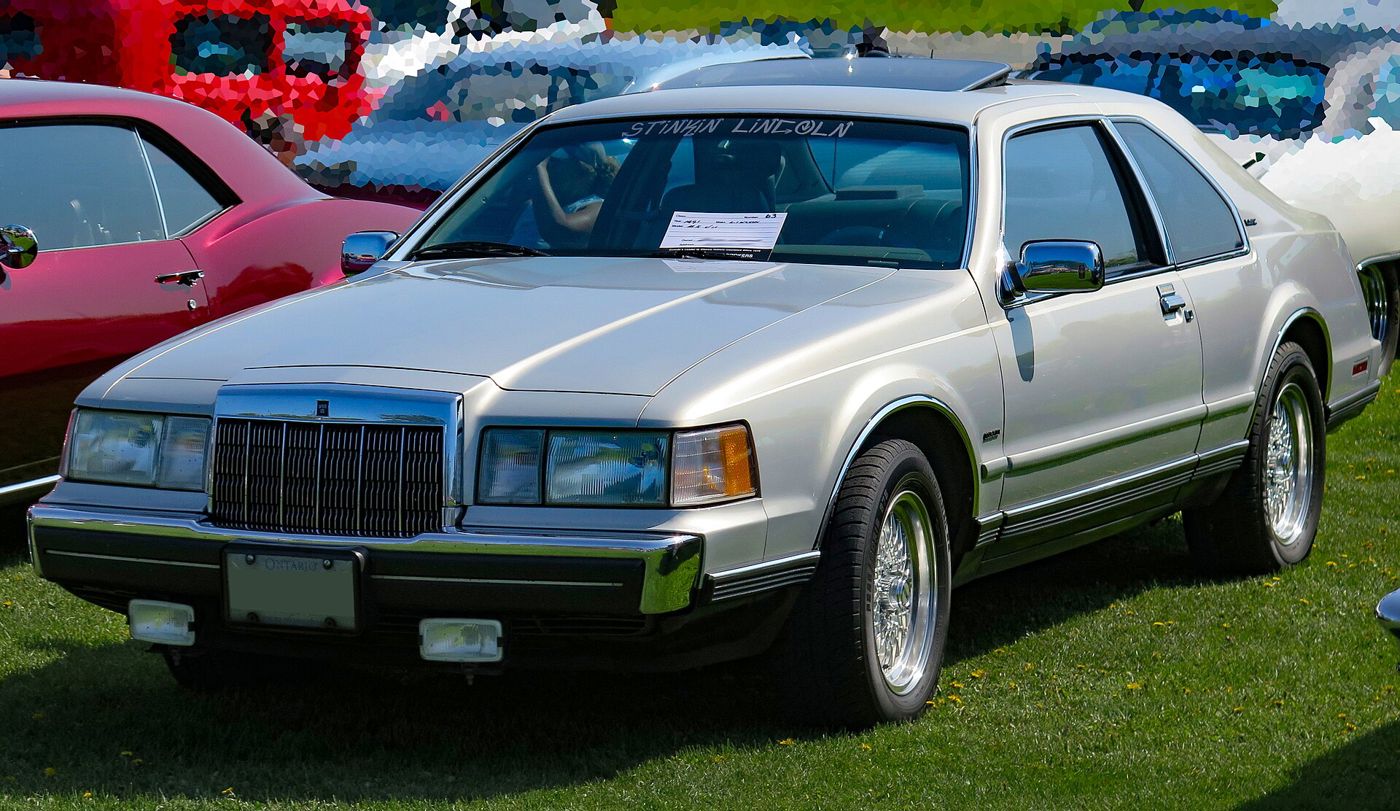Silver-grey 1991 Lincoln Mark VII LSC coupe at a cruise-in, front three-quarter view, with BBS-style wheels and a dealer name across the windshield