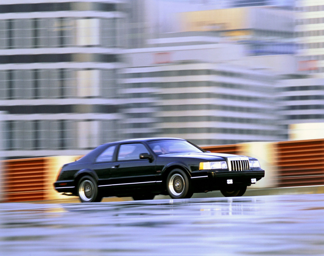 Black 1992 Lincoln Mark VII coupe with BBS-style wheels, driving past blurred buildings in a Ford press photo