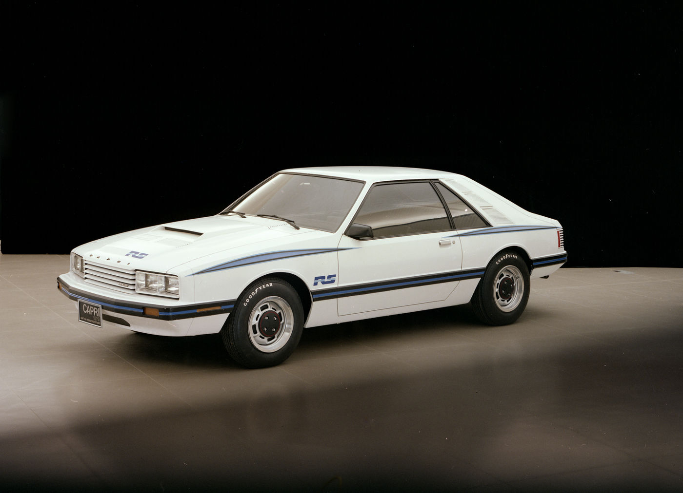 White 1979 Mercury Capri RS hatchback with blue body stripes, RS decals and a hood scoop, front three-quarter studio view on a black backdrop