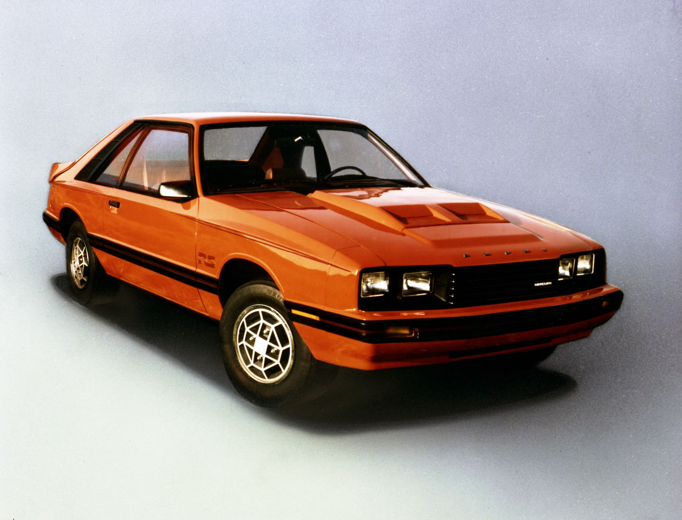 Orange 1981 Mercury Capri RS hatchback with a black lower band, hood scoop and louvered alloy wheels, front three-quarter studio view