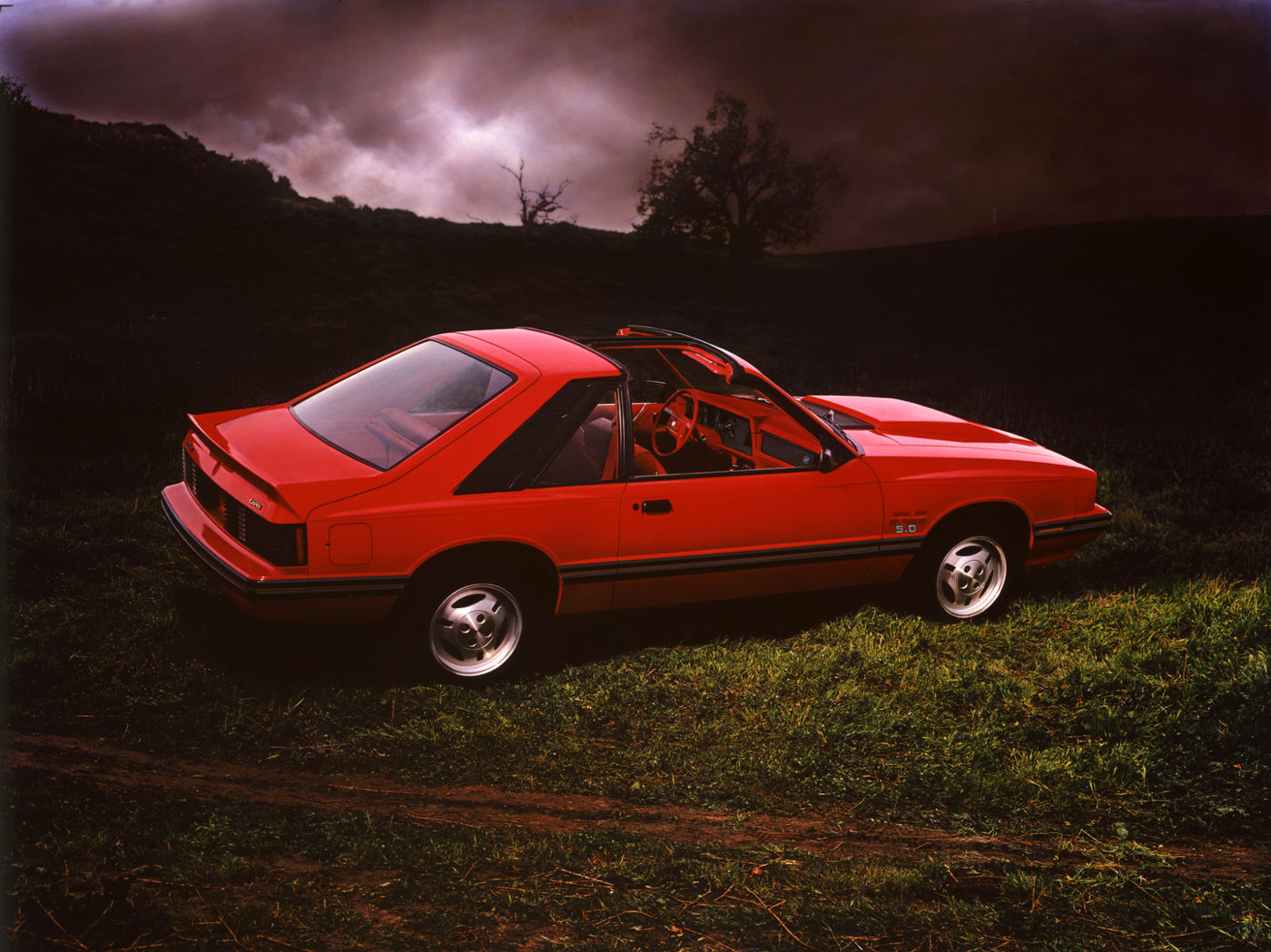 Red 1982 Mercury Capri RS hatchback with its roof panels open, rear three-quarter view on a grassy hillside under a stormy sky, 5.0 badge on the front fender