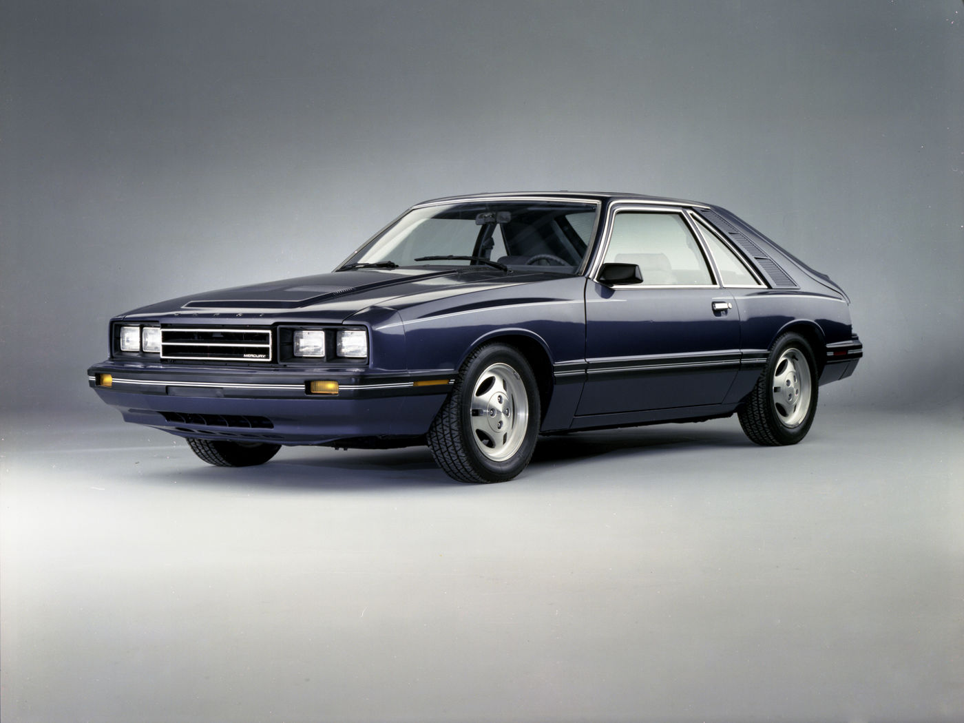 Dark blue 1983 Mercury Capri hatchback in a studio, front three-quarter view, showing the four-headlight nose and the curved rear hatch glass