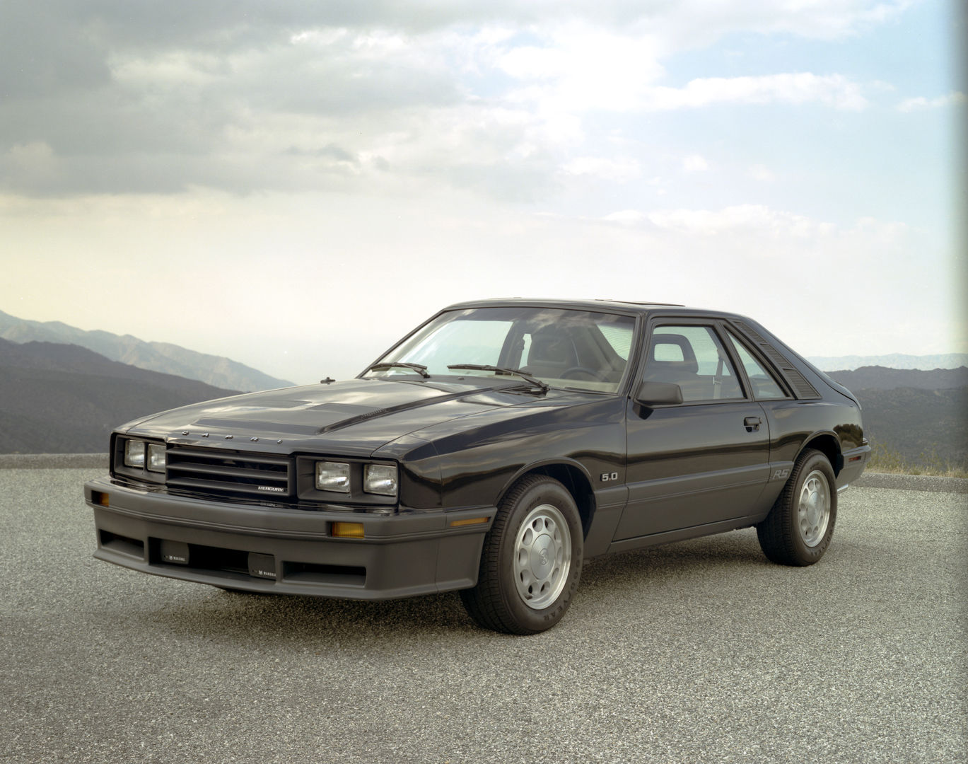 Black 1985 Mercury Capri RS hatchback with a 5.0 fender badge and hood bulge, front three-quarter view on a gravel overlook with mountains behind