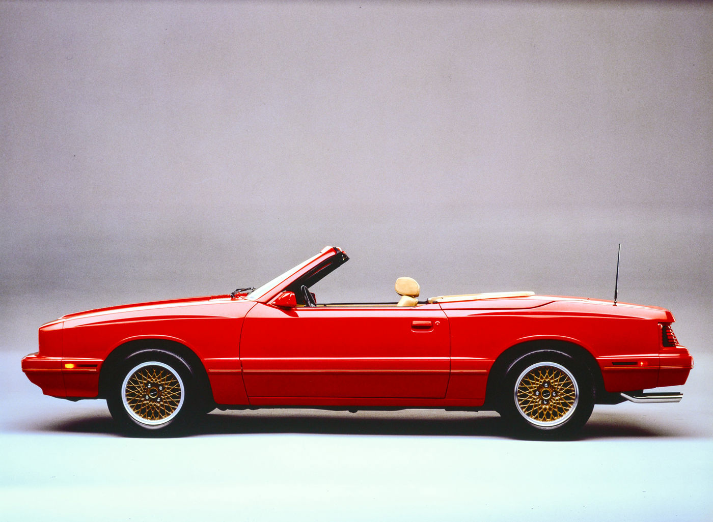 Red 1986 Mercury Capri convertible in full side profile in a studio, top down, with gold basket-weave wheels and a body-color lower valance