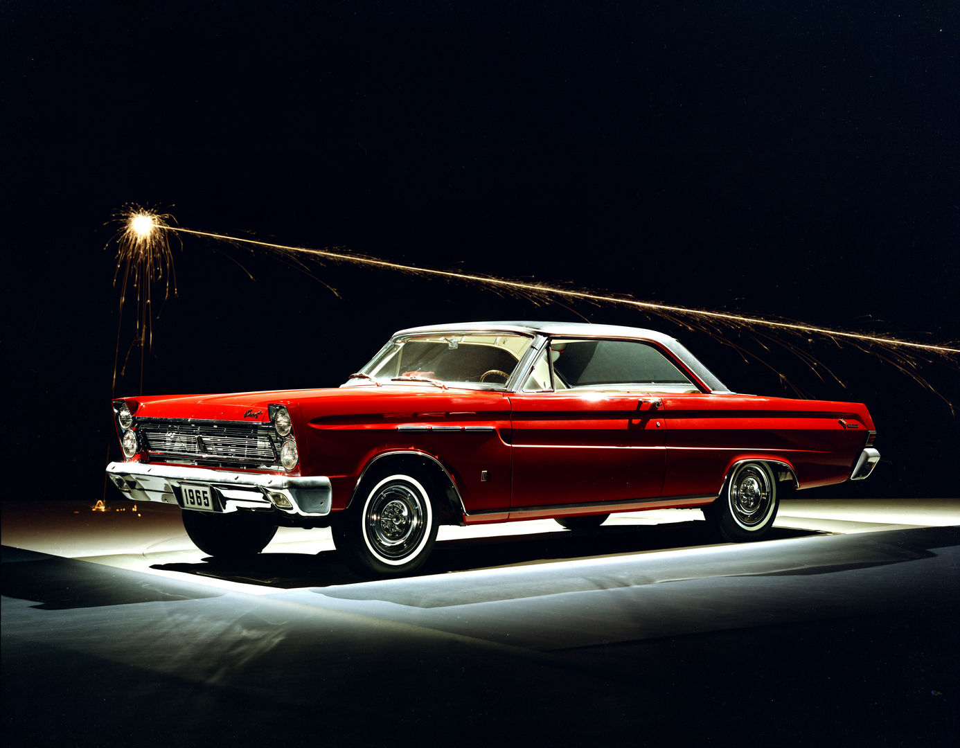 Red 1965 Mercury Comet Cyclone two-door hardtop with a white roof, lit from below on a black stage with a firework streak overhead, showing the stacked quad headlights