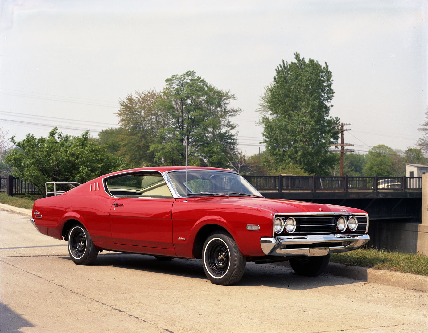 Red 1968 Mercury Cyclone fastback on a concrete apron by a small bridge, front three-quarter view, with roof-pillar vents and a Cyclone script on the rear fender