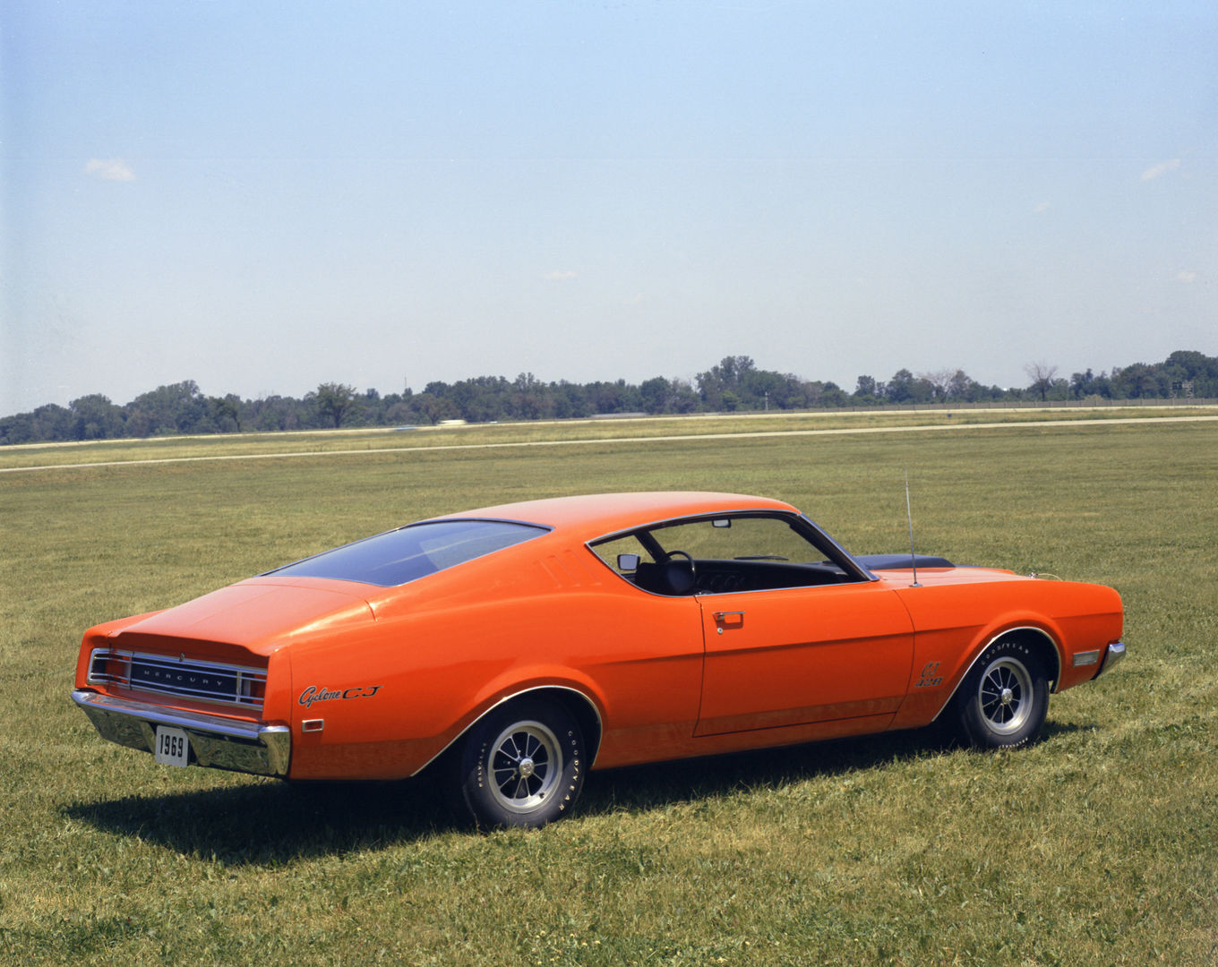 Orange 1969 Mercury Cyclone CJ fastback on a grass field, rear three-quarter view, with Cyclone CJ script on the rear fender and styled wheels