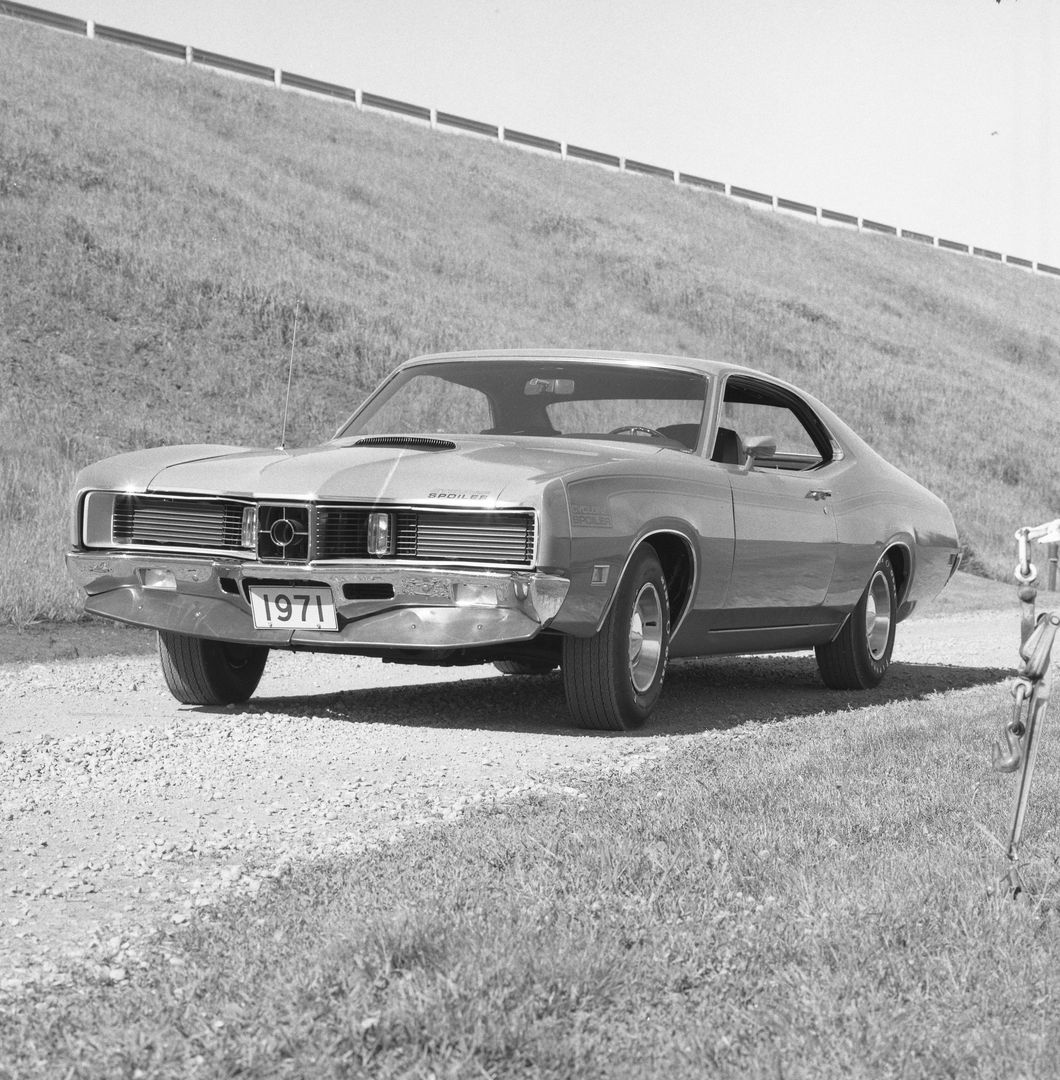 Black-and-white photo of a 1971 Mercury Cyclone Spoiler two-door hardtop on a gravel road below a grassy embankment, front three-quarter view, with Spoiler lettering on the hood