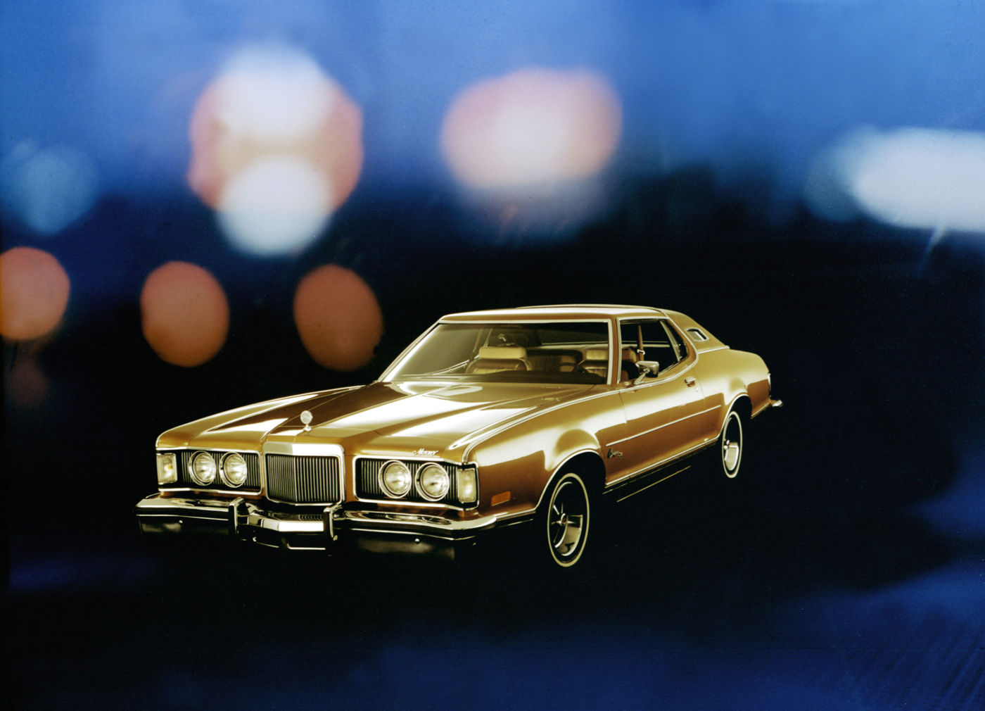 Gold 1975 Mercury Cougar two-door against a dark blue backdrop with blurred colored lights, front three-quarter view