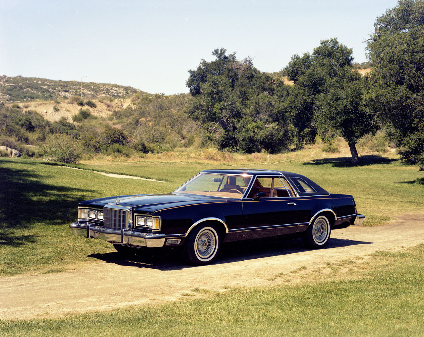 Dark blue 1977 Mercury Cougar two-door on a dirt track in rolling country, front three-quarter view, showing the upright grille and hidden headlights