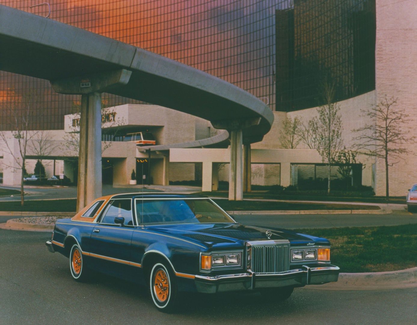 Dark blue 1979 Mercury Cougar XR-7 with gold stripes and gold wheels in front of a hotel with a curved elevated walkway